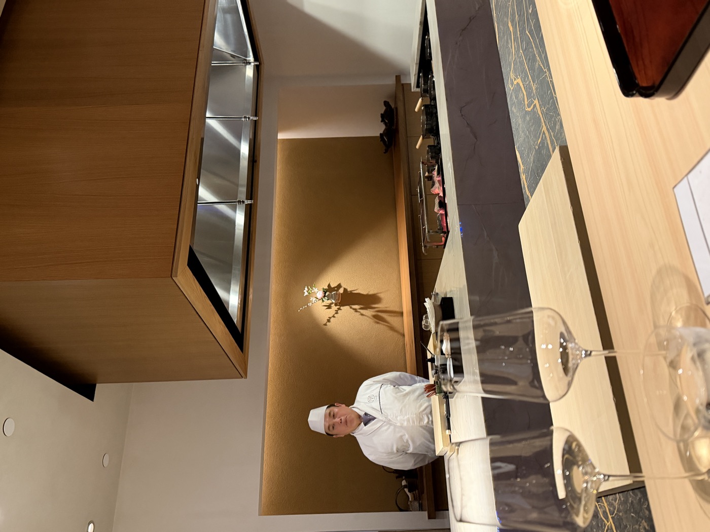 A bare hinoki-wood chef's counter, a single chef in white standing behind it beside an ikebana arrangement