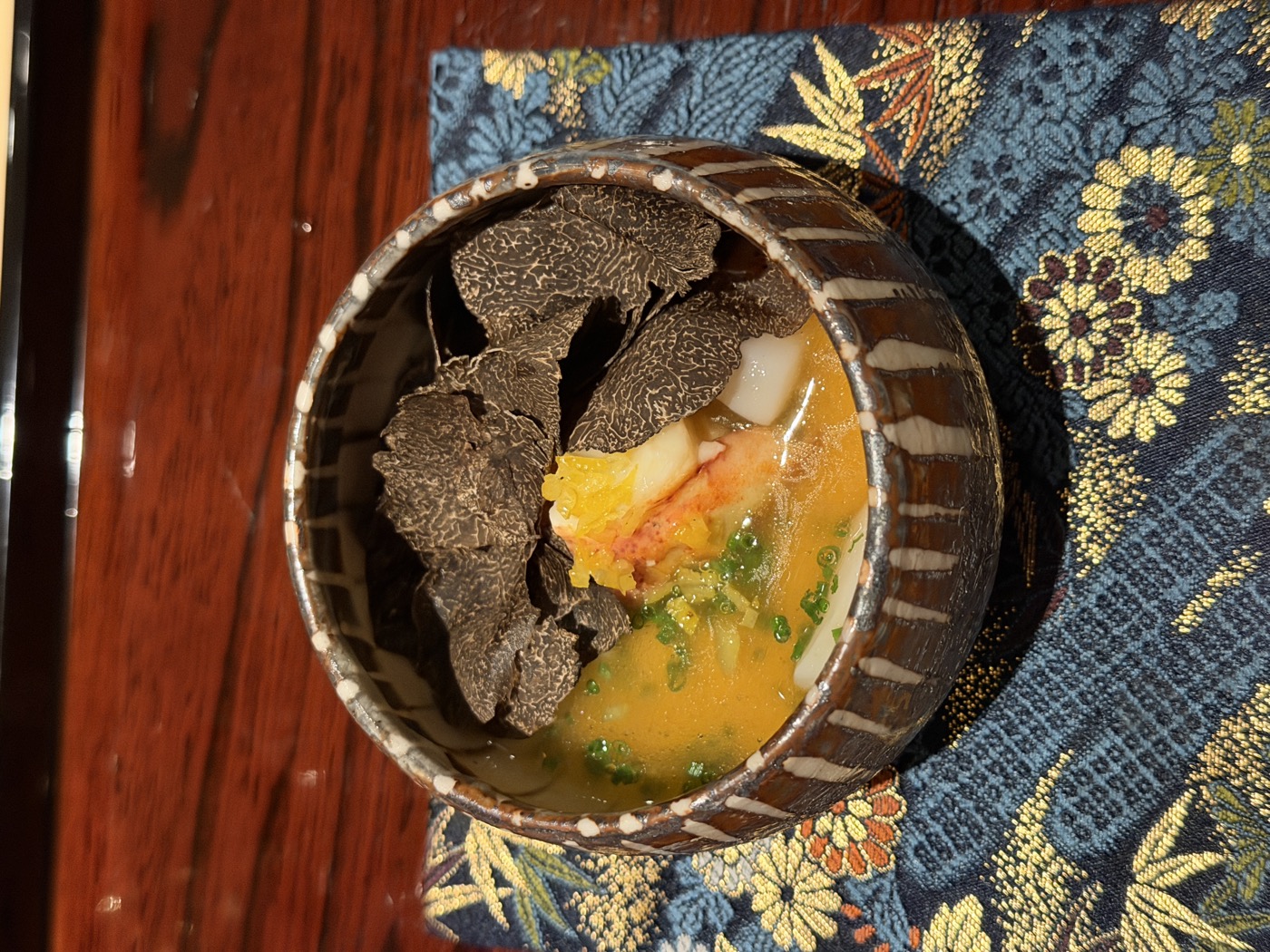 A rustic bowl on a gold-and-blue chrysanthemum cloth, a warm savory custard blanketed in shaved black truffle