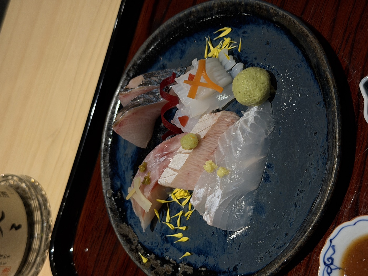 A dark-blue glazed plate of sashimi — white fish and yellowtail — with chrysanthemum petals and a ball of fresh wasabi