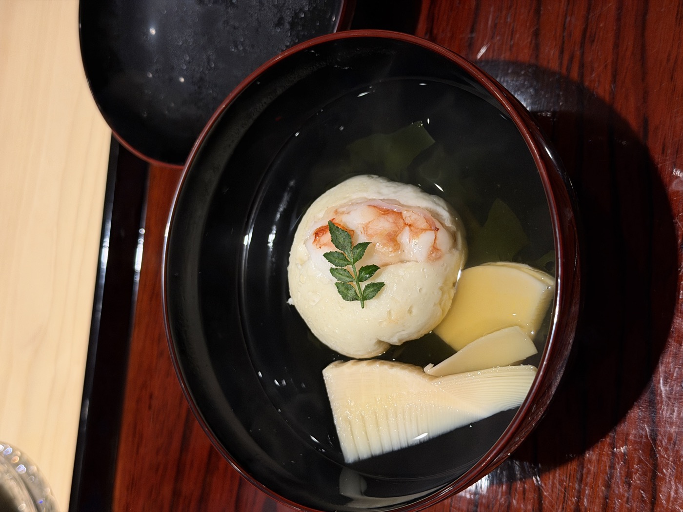 A lacquer bowl of clear broth with a white shrimp dumpling, a slice of bamboo shoot and a sprig of kinome