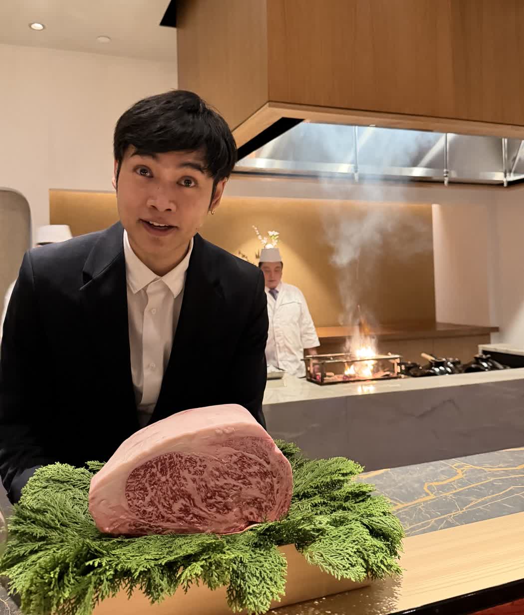 A whole block of marbled wagyu beef held up on a bed of hinoki greenery, the grill smoking behind