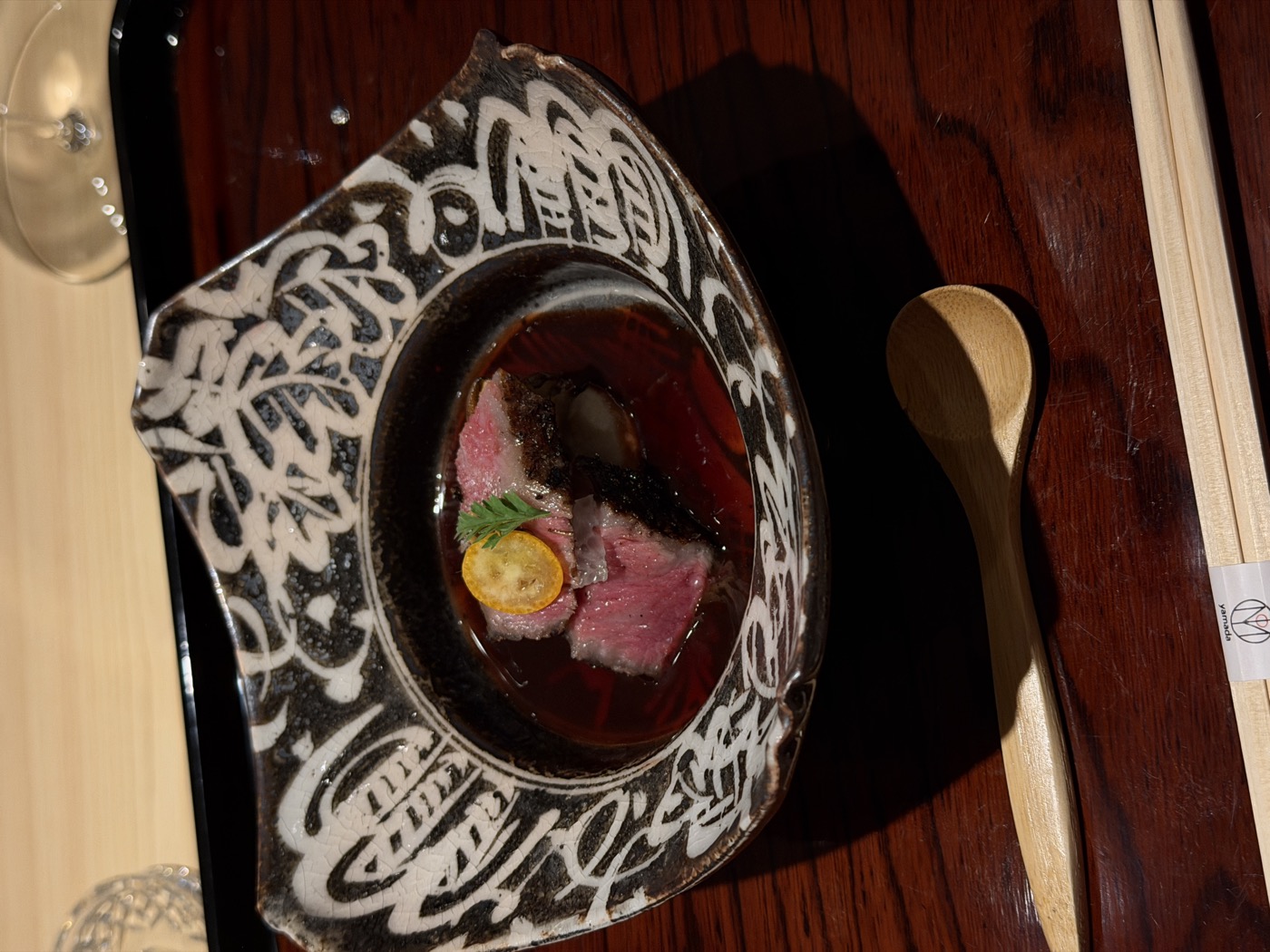Blushing slices of wagyu in a glossy dark broth in a patterned bowl, with a coin of kumquat