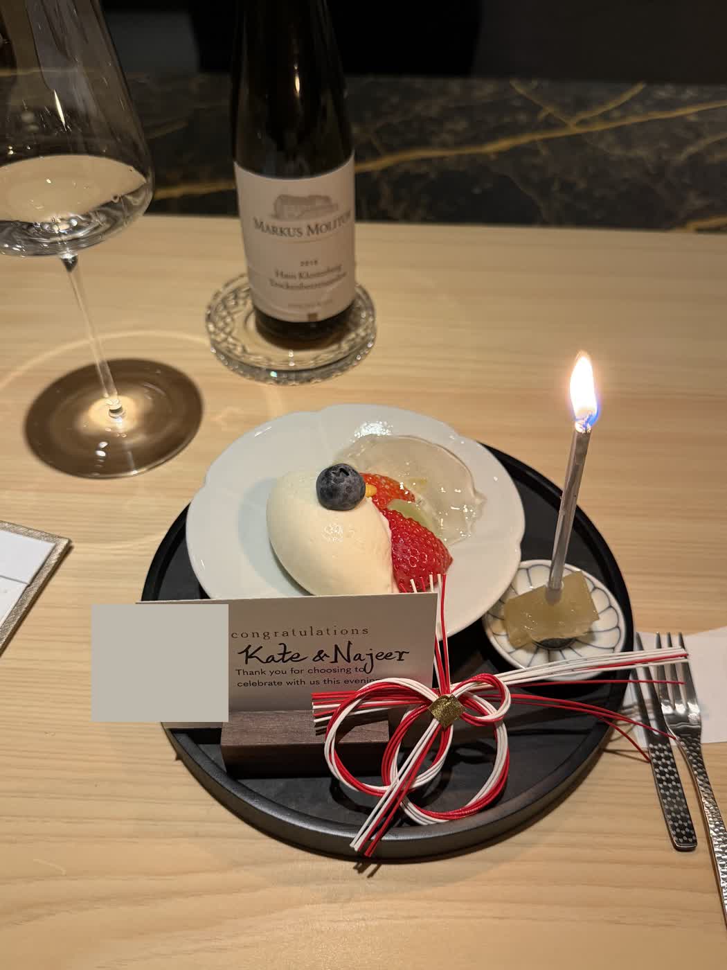 A white plate of amazake ice cream with strawberry and blueberry, beside a lit candle and a note reading congratulations Kate and Najeer