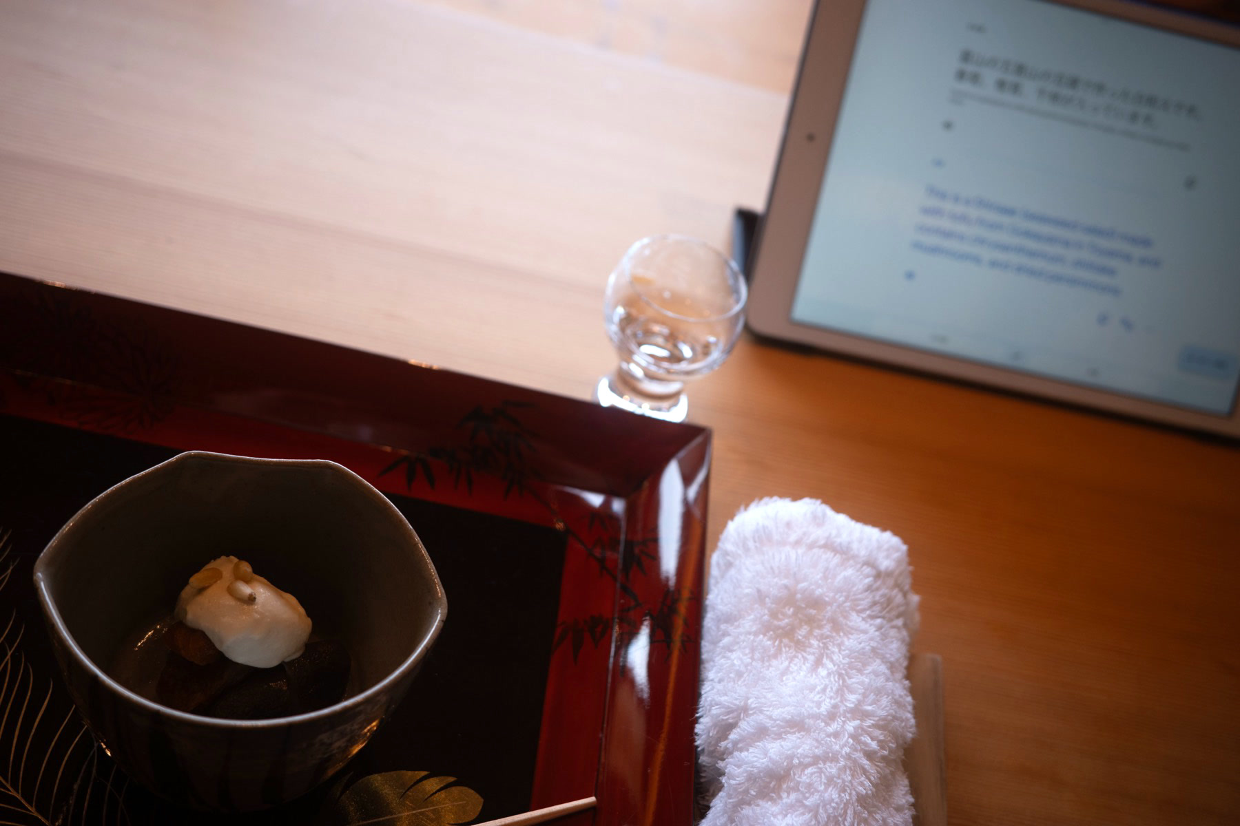 A small bowl holding a pale, custardy bite on a dark lacquer tray, beside a glass of sake and a folded towel