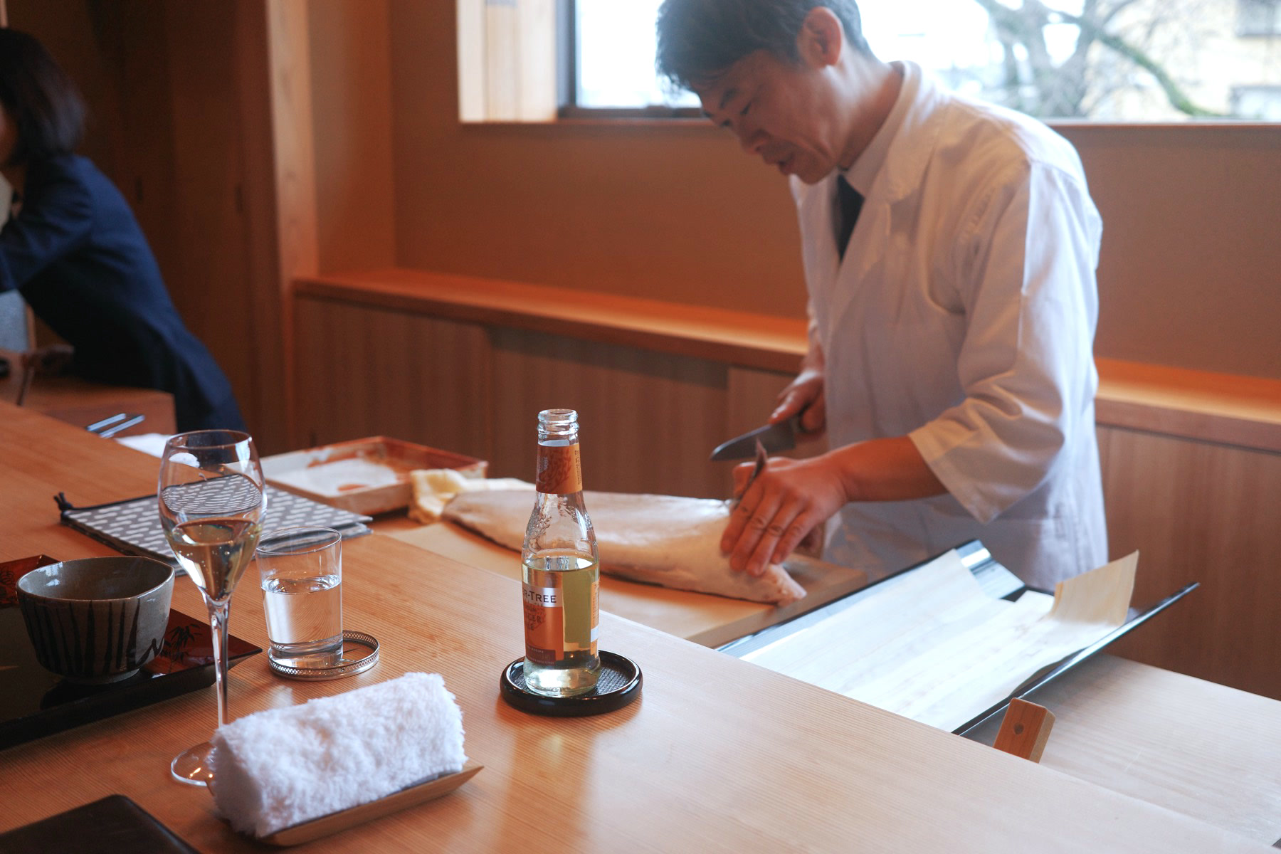 The chef slicing fish behind the counter, our glasses and a bottle in the foreground