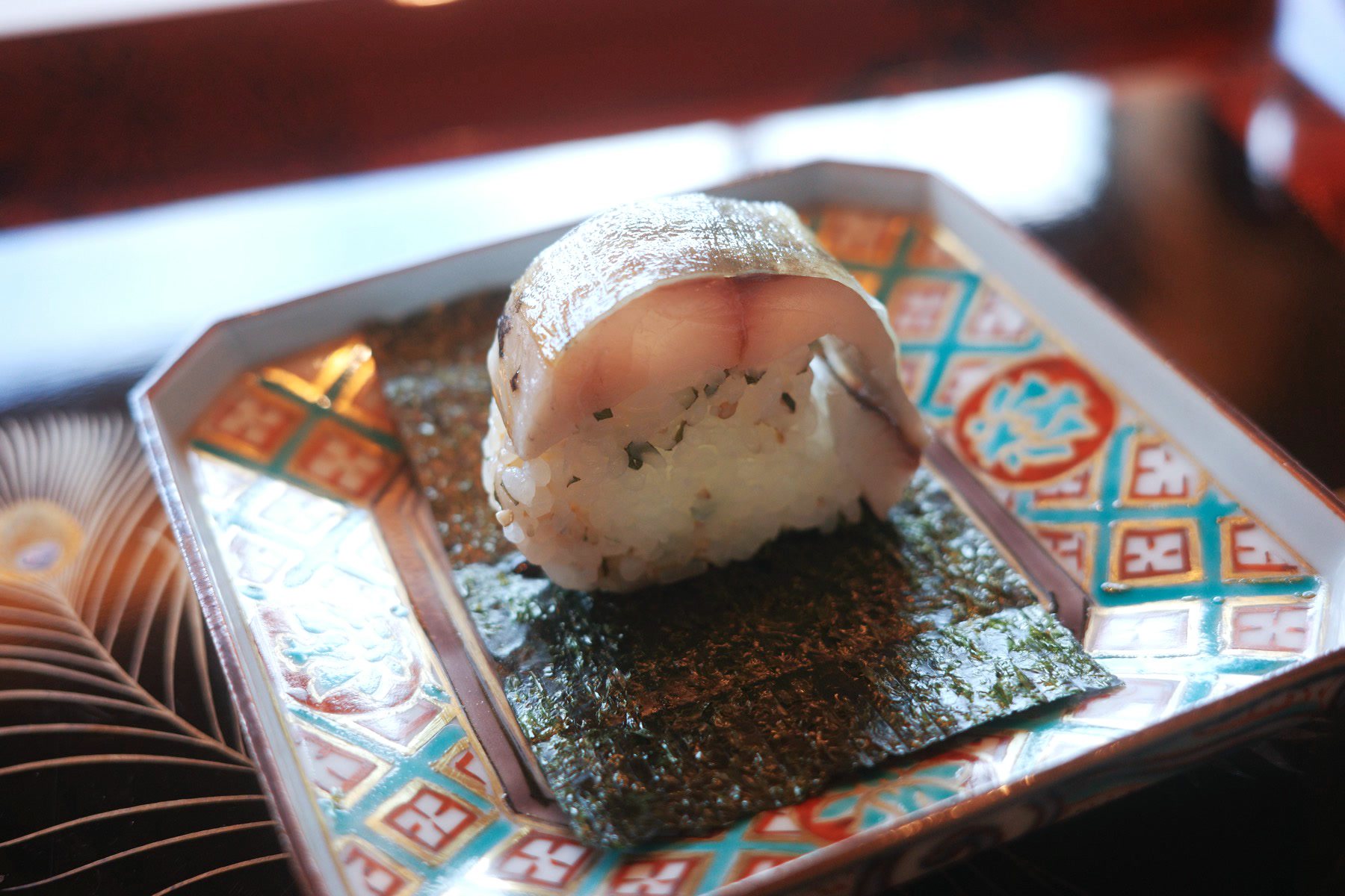 A block of pressed horse-mackerel sushi resting on a sheet of nori, on a patterned plate