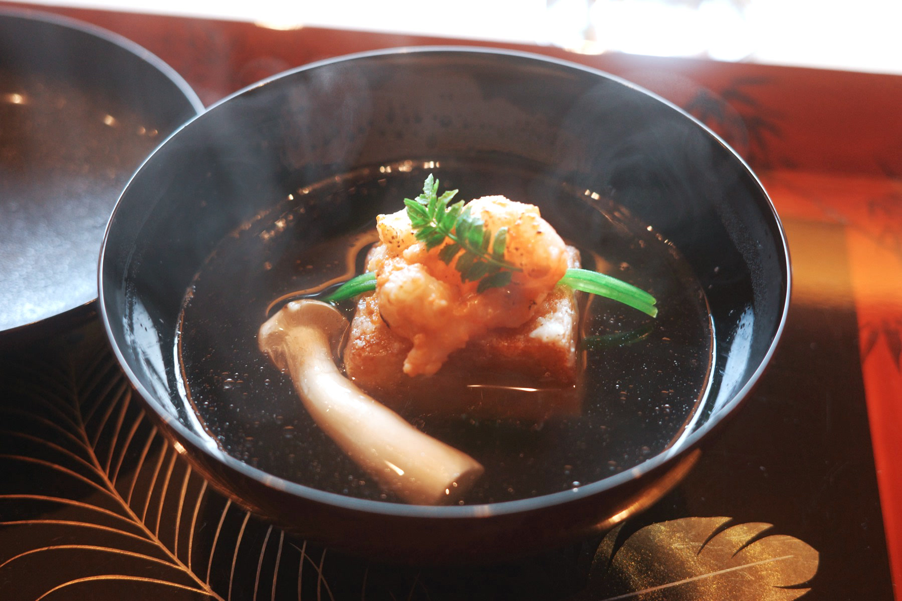 A dark lacquer bowl of clear broth with a fried taro dumpling topped with shrimp and a herb sprig