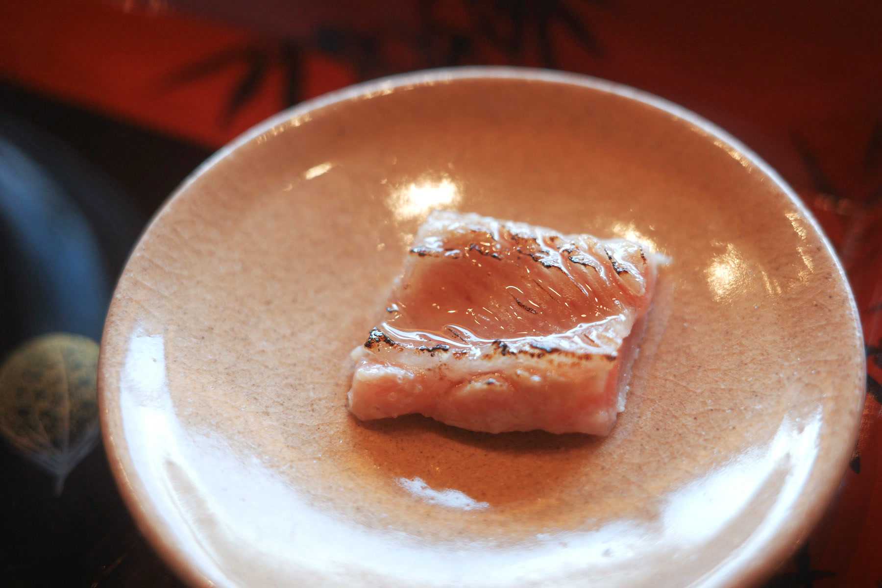A single slice of fatty fish, lightly seared along one edge, on a plain ceramic plate