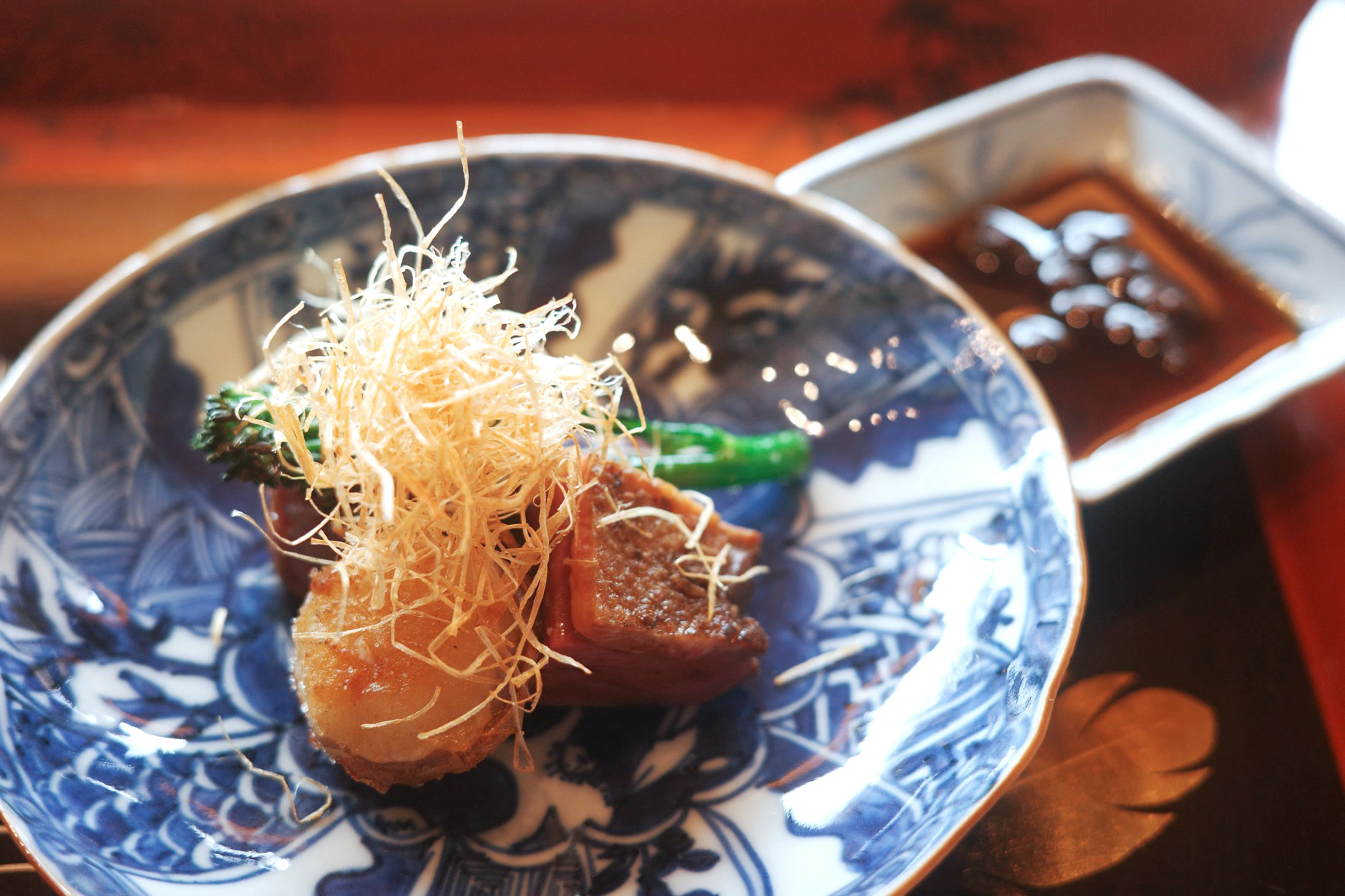 Slices of seared duck on a blue-and-white plate, topped with crisp-fried threads and broccolini, with a dish of sauce