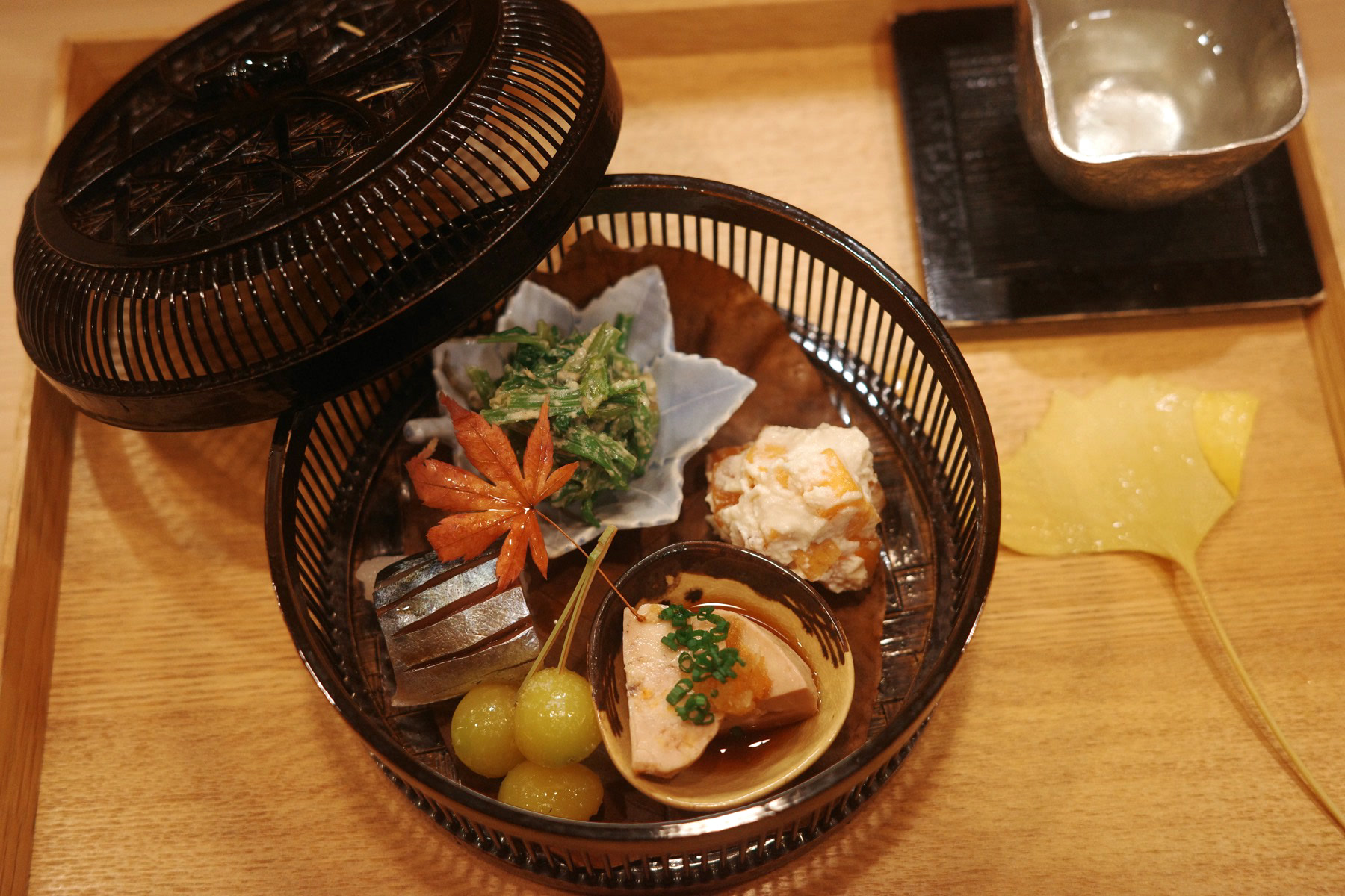 A woven bamboo basket of small seasonal bites, beside a silver sake cup and a ginkgo leaf