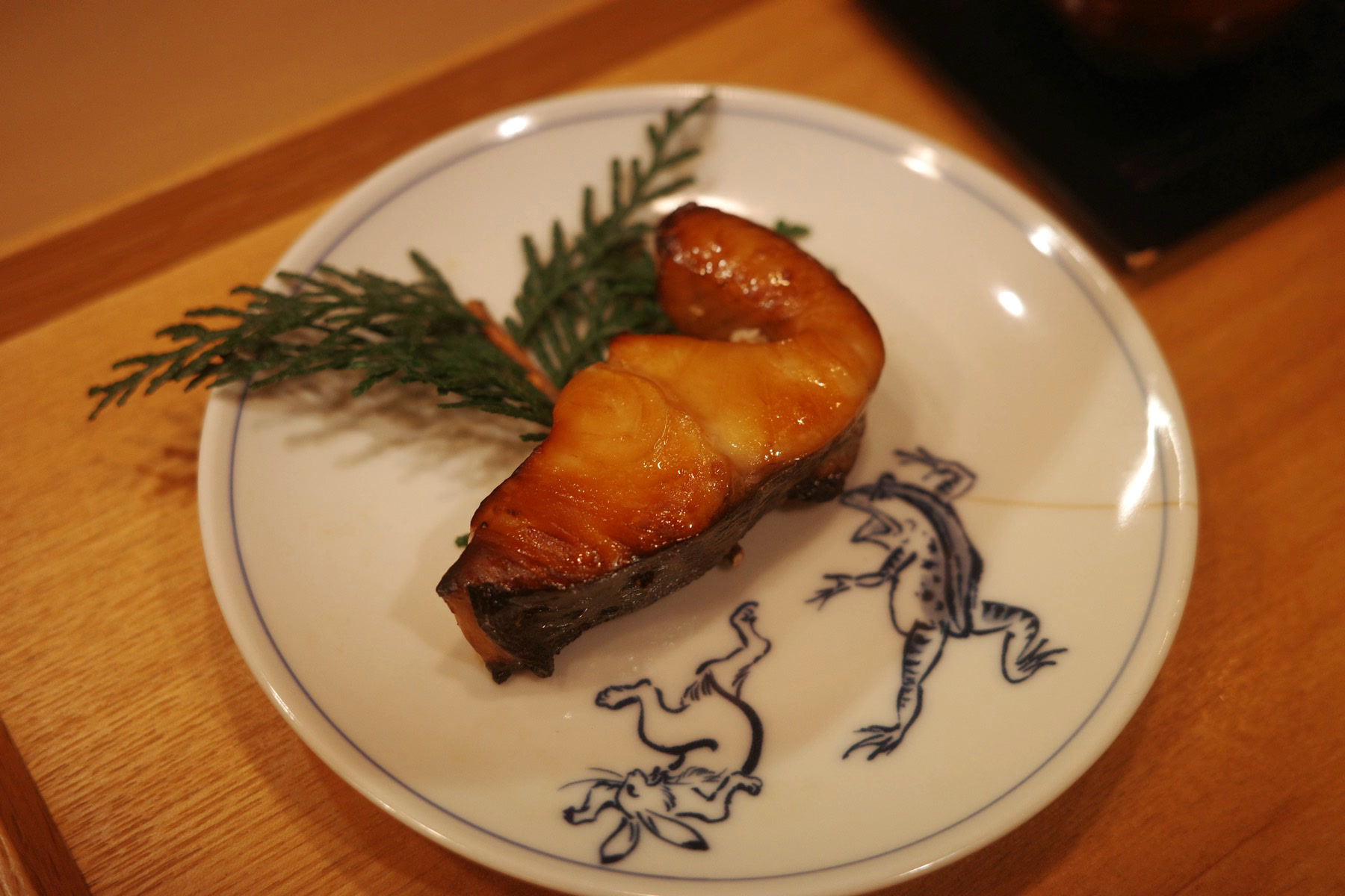 A glazed grilled fish fillet on a frog-and-rabbit painted plate